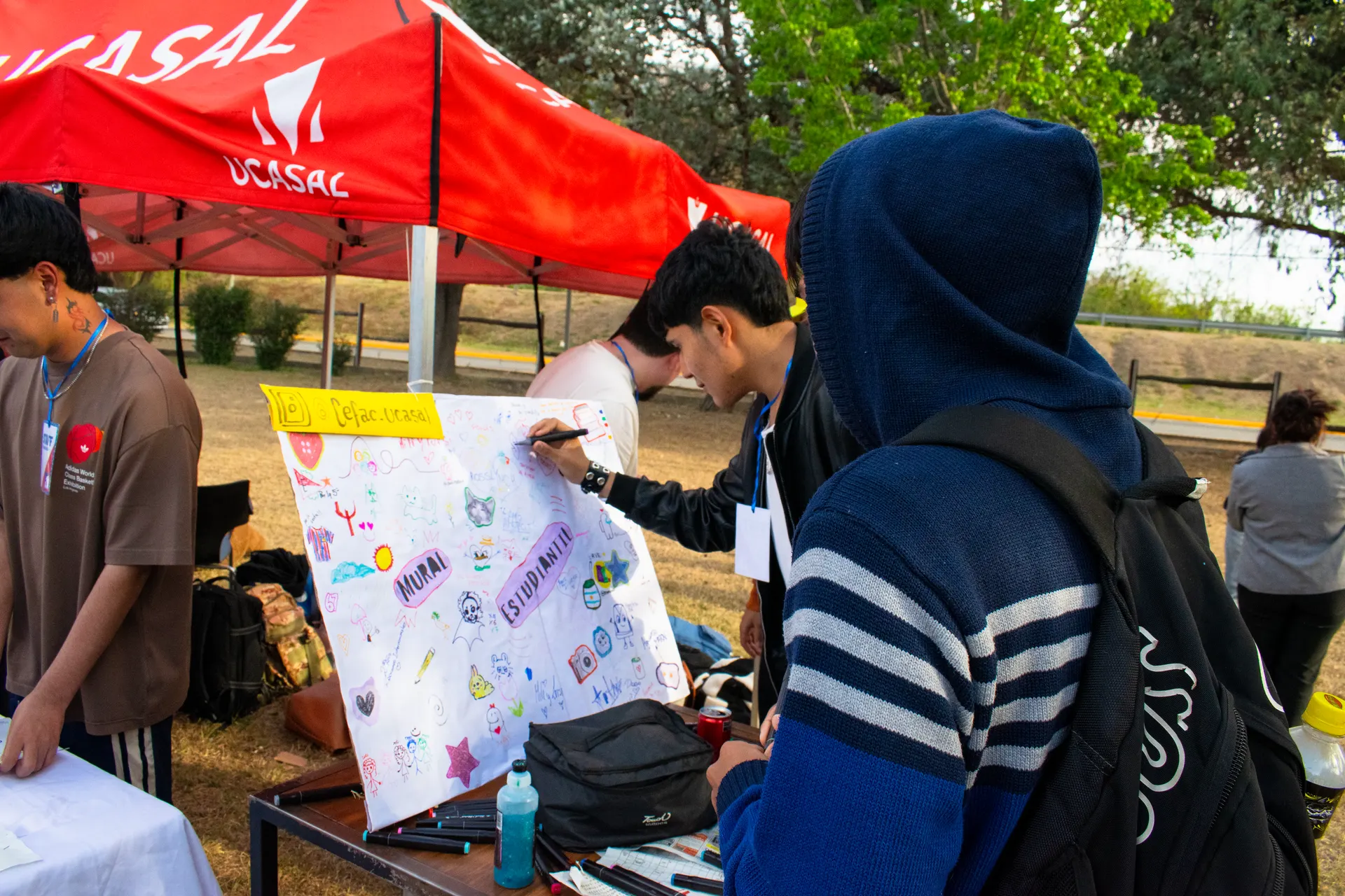 Estudiantes dibujan sobre un mural de tela con la palabra «Estudiante» bajo una carpa roja.